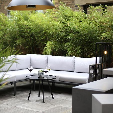 Middle Eight - Covent Garden - Preferred Hotels And Resorts London Ngoại thất bức ảnh The photo shows an outdoor seating area featuring a modern gray sectional sofa with plush cushions. In front of the sofa is a small, round black table with two glasses of wine on it, accompanied by a decorative plant or vase. Surrounding the area are